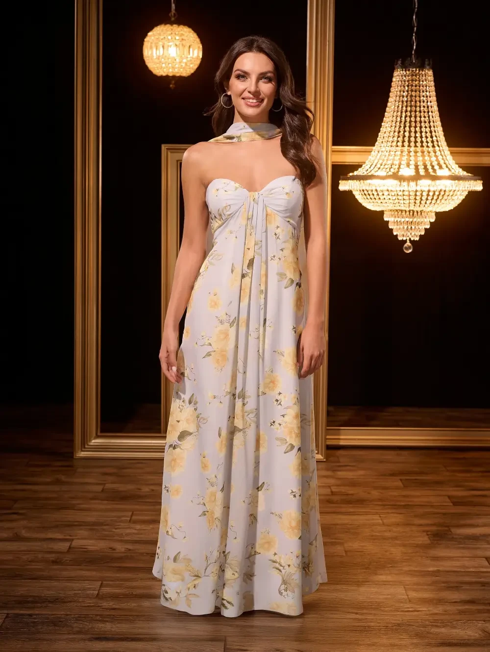 A woman wearing a floral-patterned, strapless maxi dress stands in front of elegant chandeliers and a decorative frame backdrop.