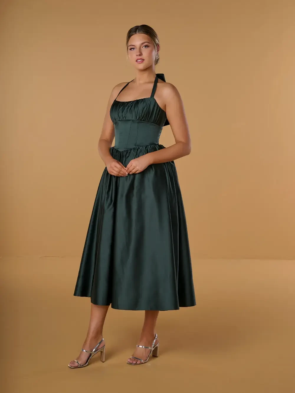 A woman in a dark green halter neck dress stands against a beige background. She has her hands clasped in front of her and wears silver strappy heels.