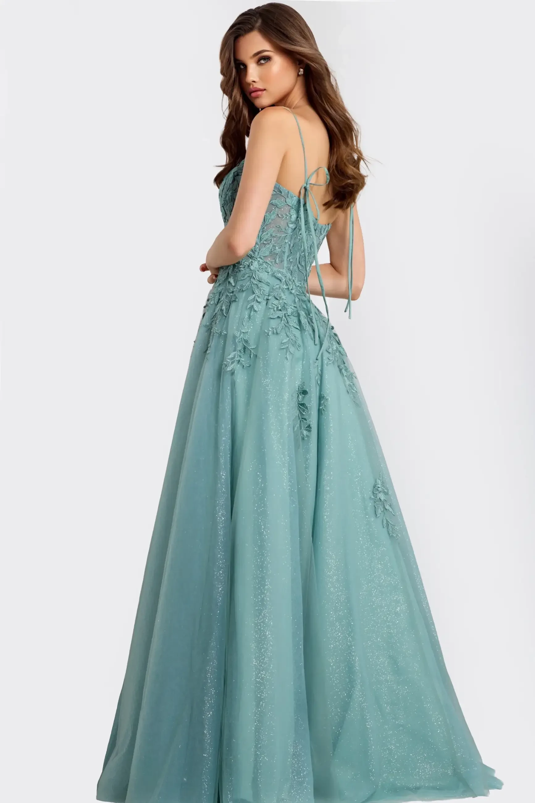 A woman in a shimmering teal gown with floral appliqu&eacute; stands sideways. The dress features thin straps and an elegant, flowing skirt, exuding a graceful and sophisticated tone.