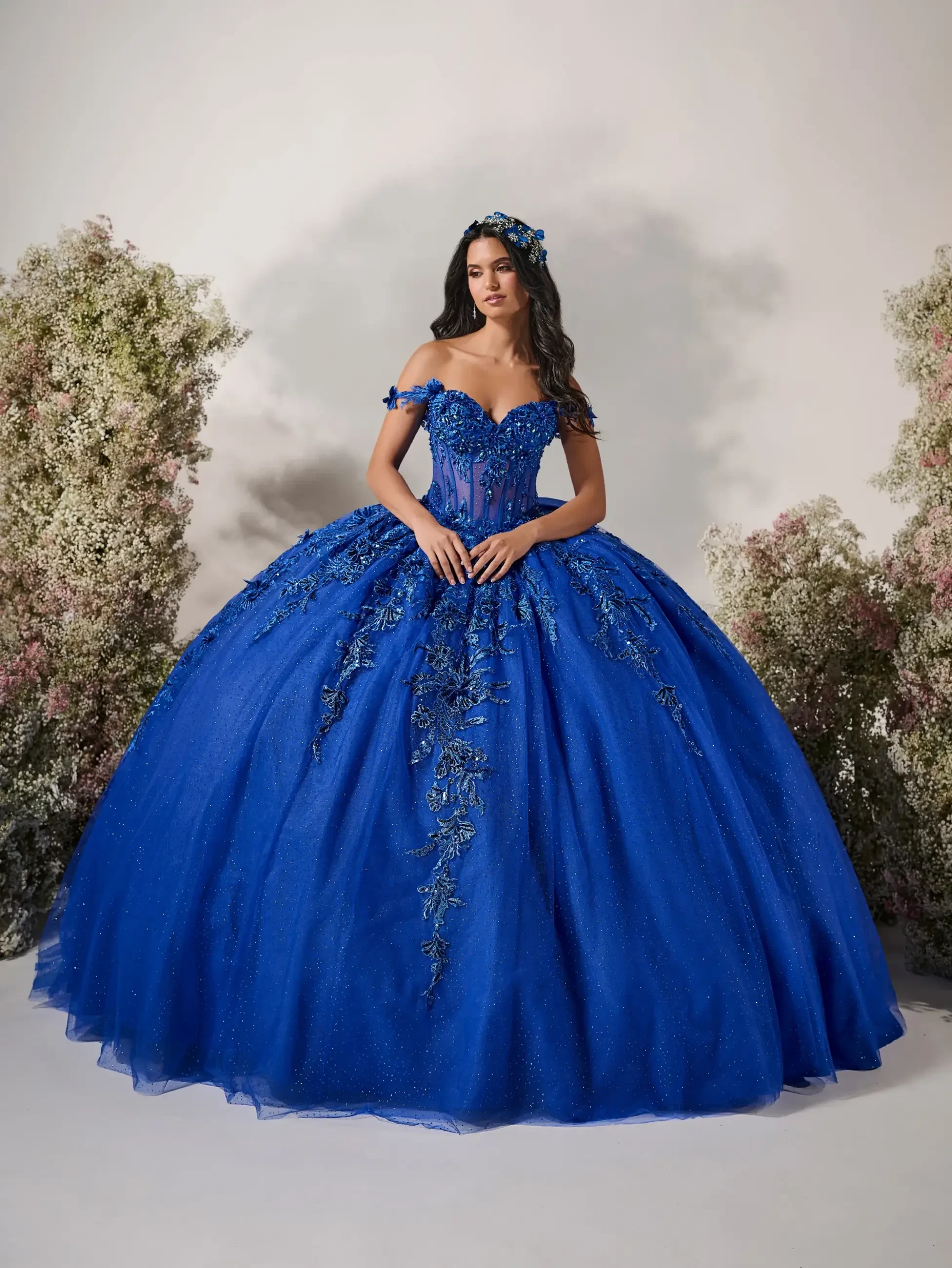 Model is wearing an off-the-shoulder sweetheart gown featuring intricate floral appliqués, a sparkling tulle skirt, and a dramatic oversized detachable bow.