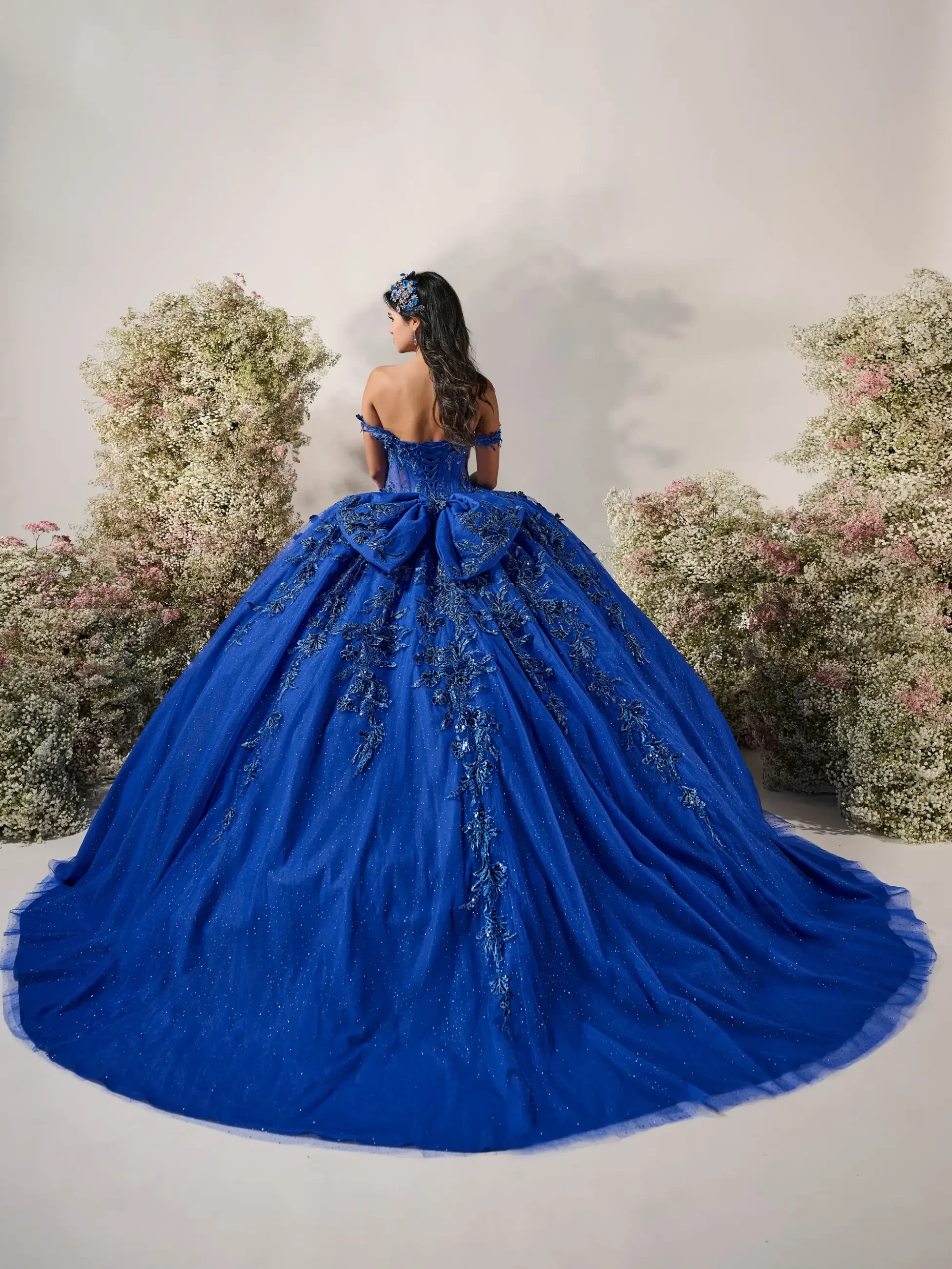 Model is wearing an off-the-shoulder sweetheart gown featuring intricate floral appliqués, a sparkling tulle skirt, and a dramatic oversized detachable bow.