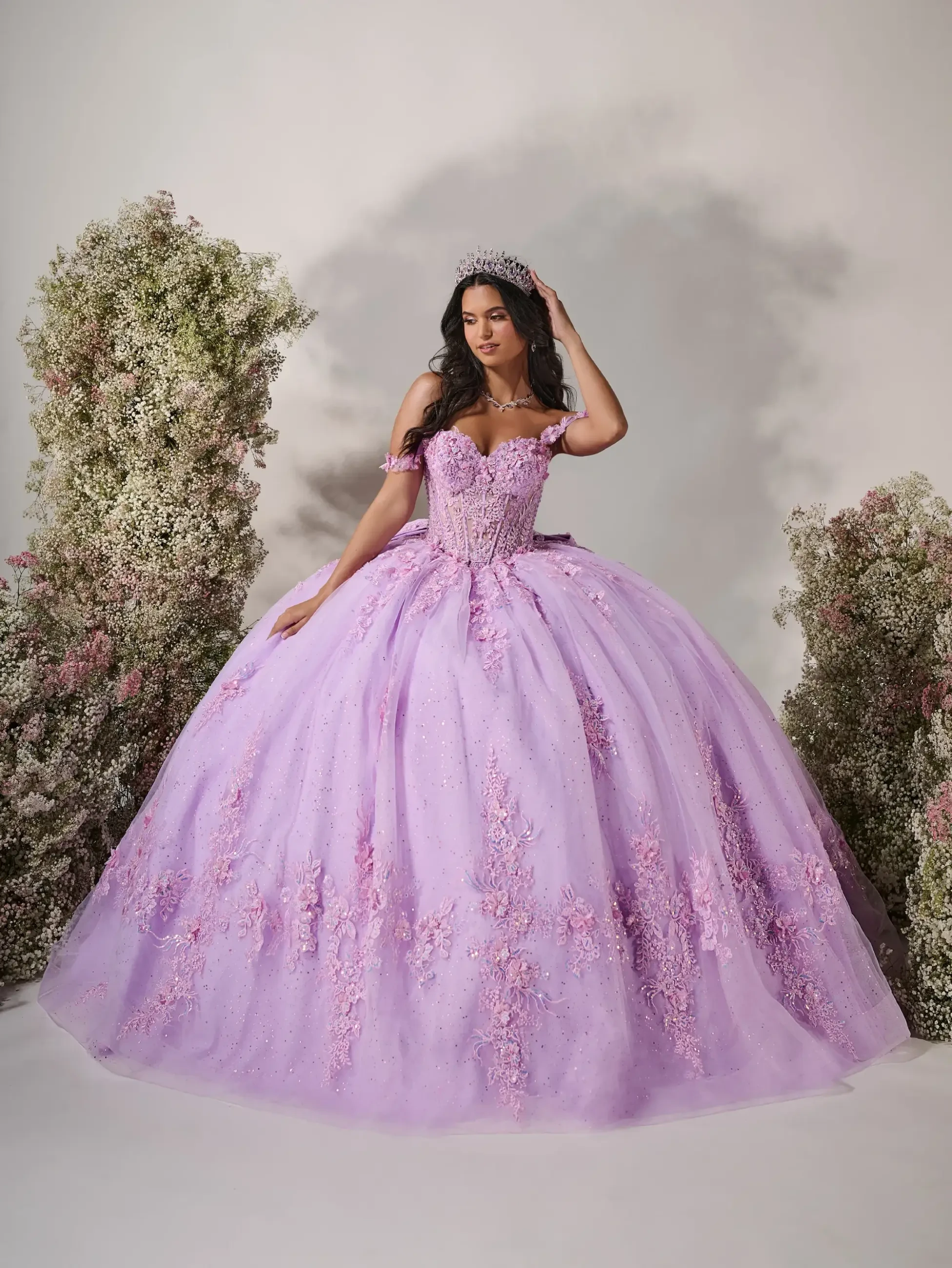 Model is wearing a sweetheart neckline gown with off-the-shoulder floral straps, a structured corset bodice, cascading embroidered appliqués, and an oversized detachable bow.