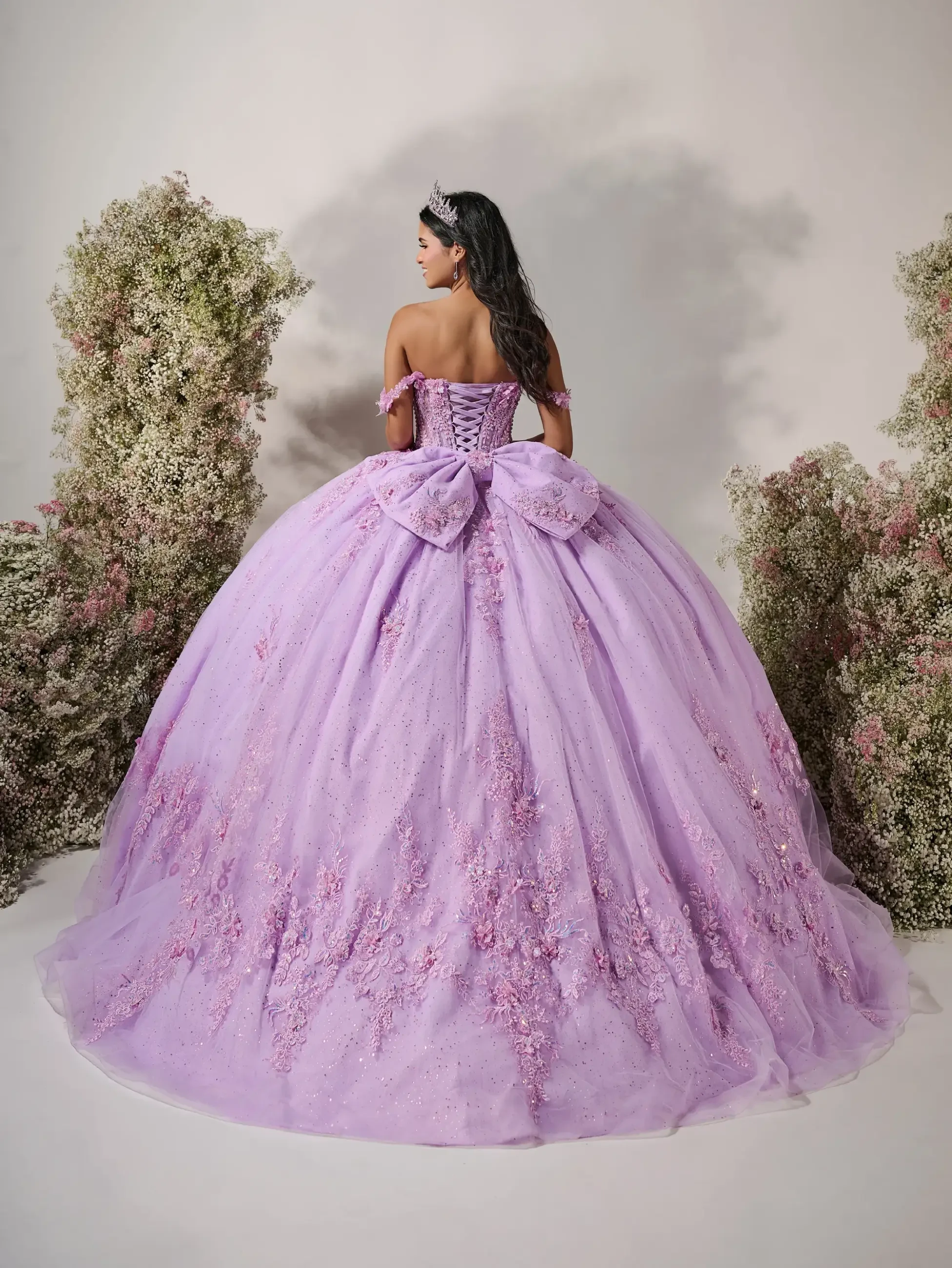 Model is wearing a sweetheart neckline gown with off-the-shoulder floral straps, a structured corset bodice, cascading embroidered appliqués, and an oversized detachable bow.