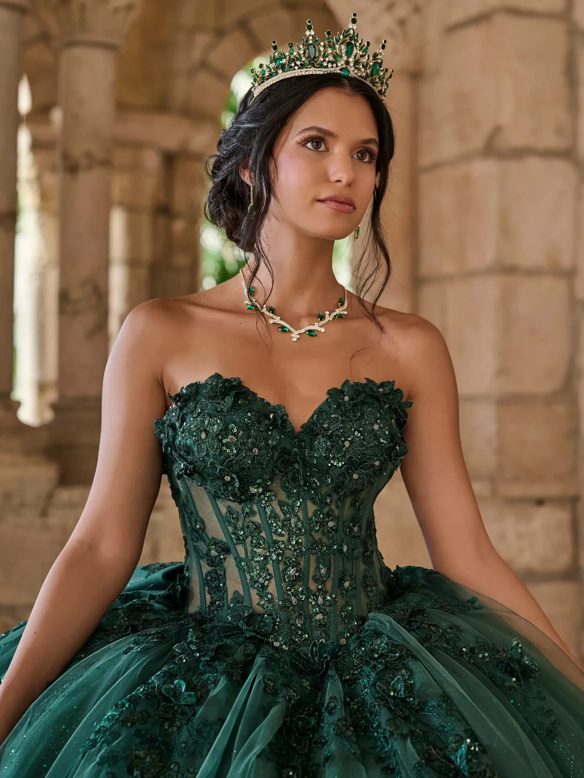 Model is wearing a strapless ball gown with a structured corset bodice adorned with large floral appliqués and a glitter tulle skirt that sparkles with every step.