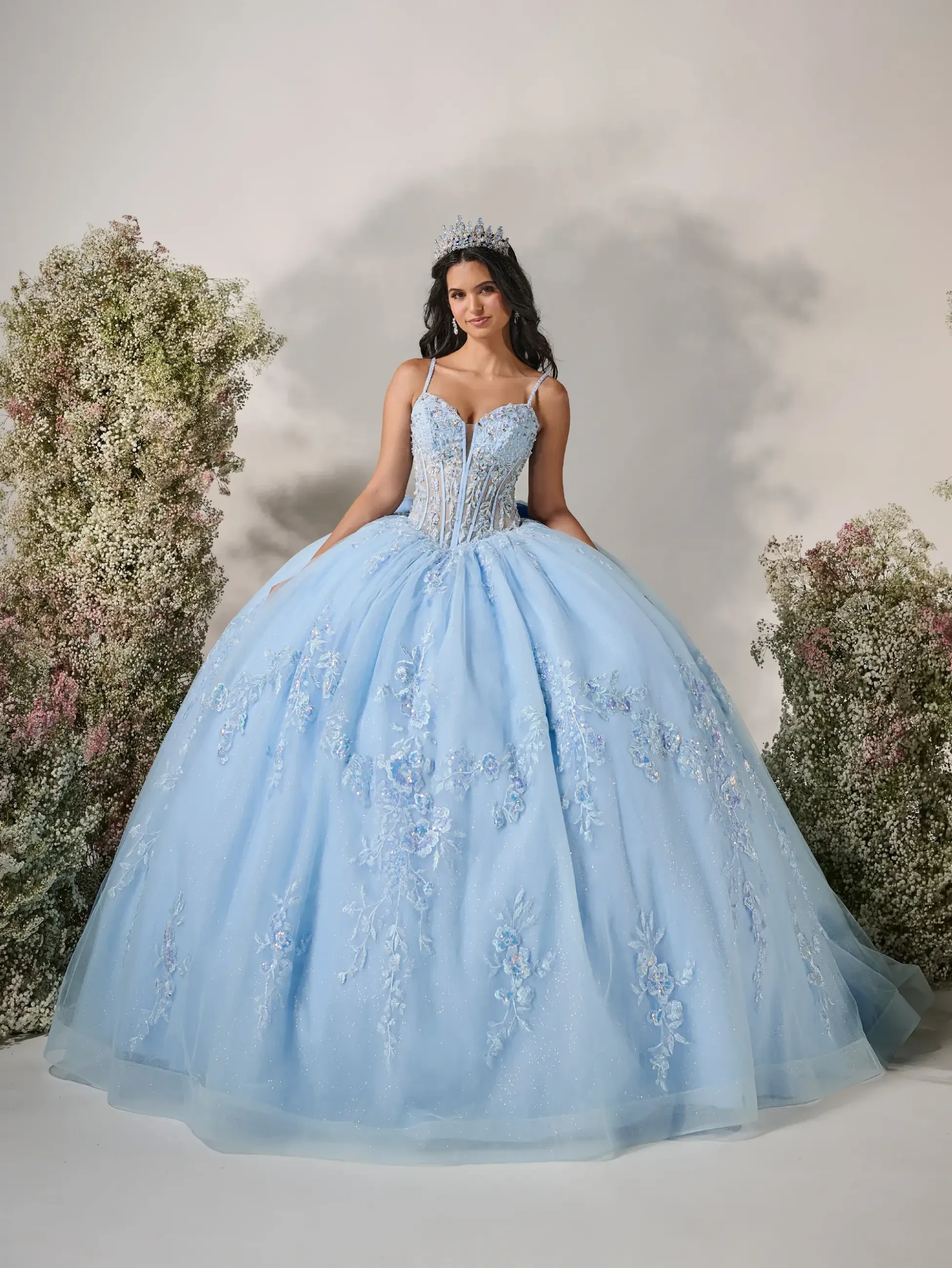 Model is wearing a romantic ball gown with a basque waist, micro plunge corset bodice, removable off-the-shoulder sleeves, and a voluminous sparkle tulle skirt adorned with delicate floral appliqués.