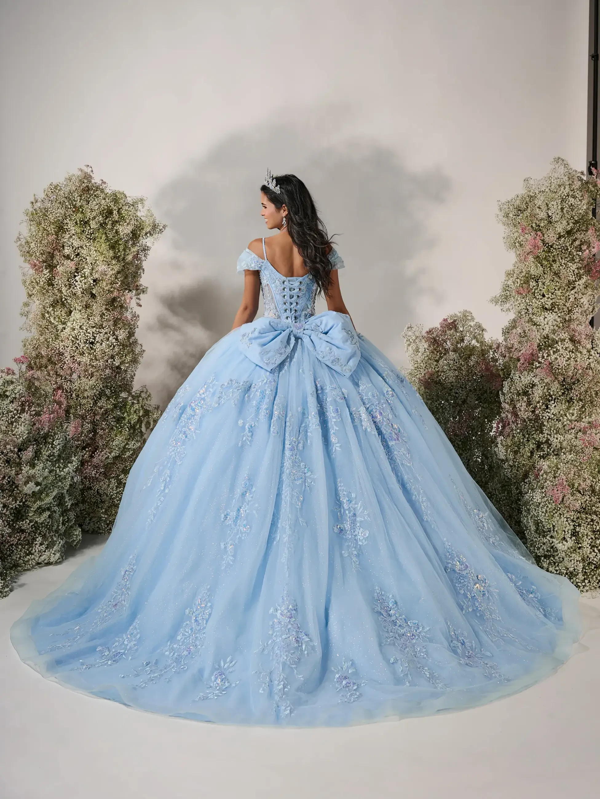 Model is wearing a romantic ball gown with a basque waist, micro plunge corset bodice, removable off-the-shoulder sleeves, and a voluminous sparkle tulle skirt adorned with delicate floral appliqués.