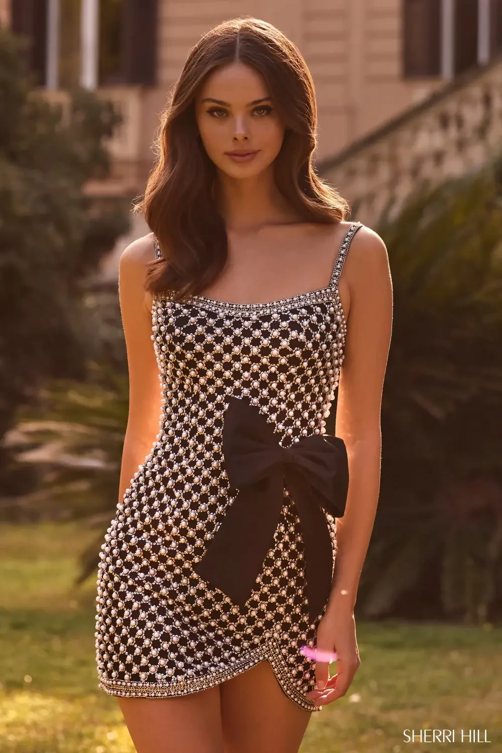 A woman stands outdoors, wearing a form-fitting, black and white dress adorned with sparkly embellishments and a large black bow at the waist.