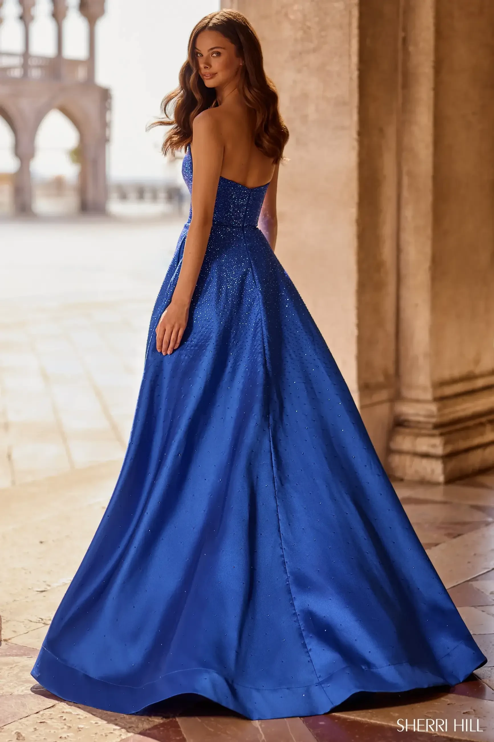 A woman in a vibrant blue, strapless gown stands on a tiled floor in a sunlit, arched hallway, exuding elegance and grace.