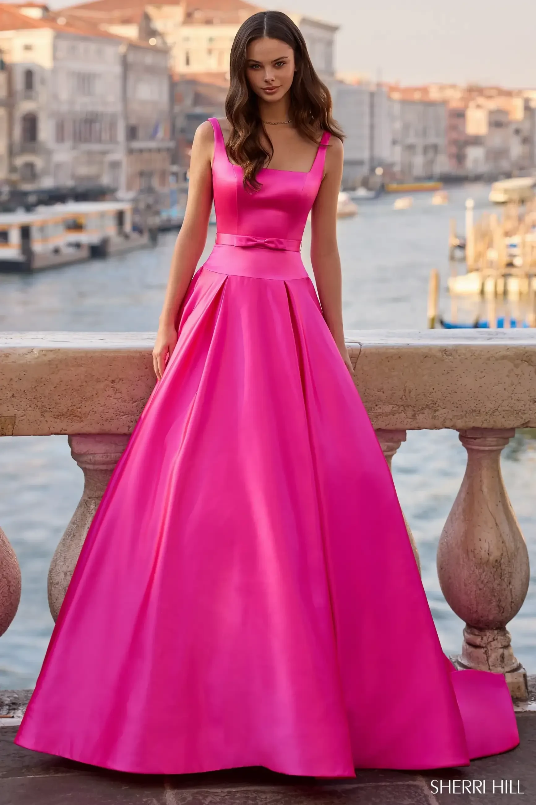 A woman in a vibrant pink ball gown with a fitted bodice stands on a balcony overlooking a canal. The scene conveys elegance and serenity.