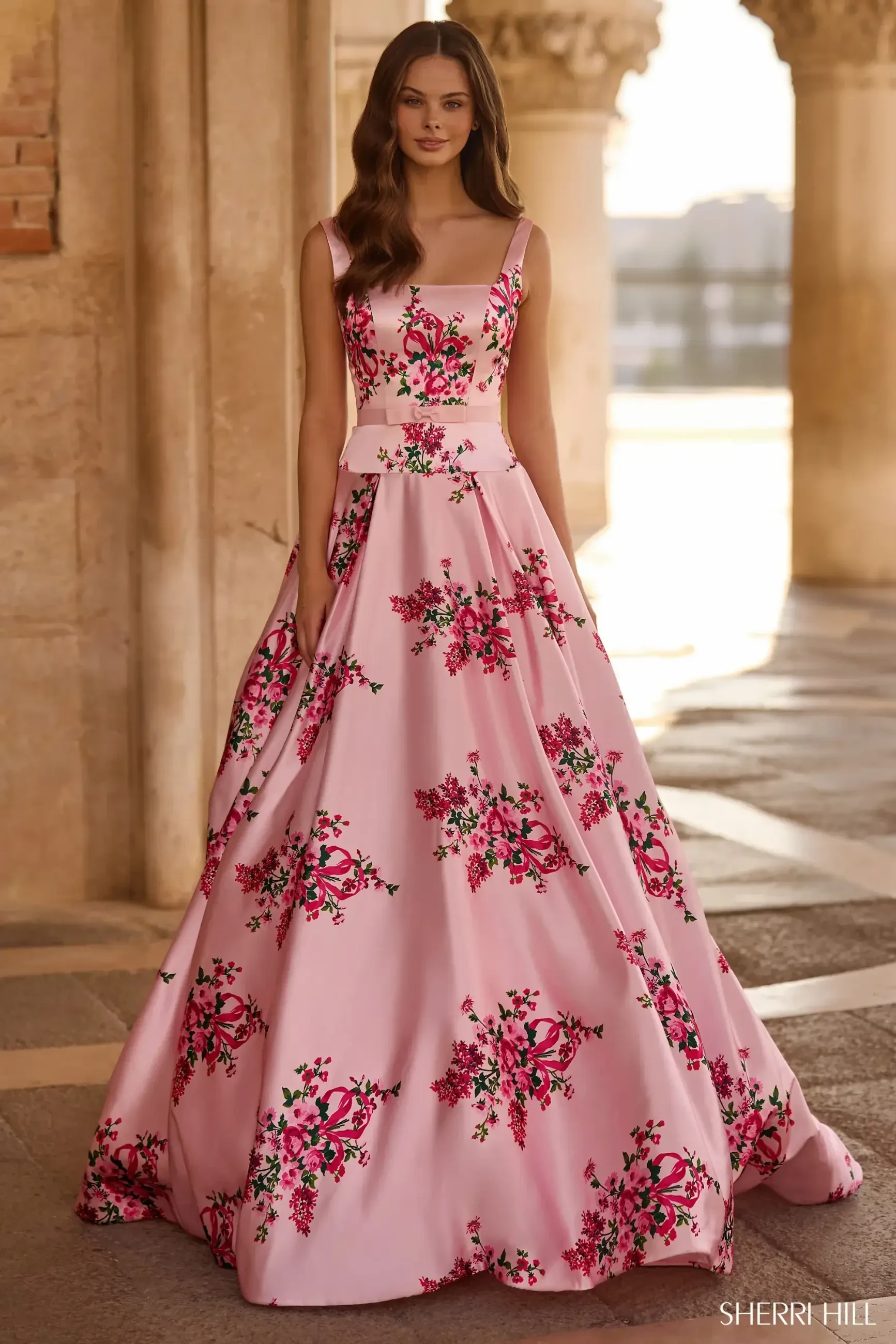 A woman in a flowing pink gown adorned with vibrant floral patterns stands elegantly in a sunlit, arch-lined hallway. The atmosphere is serene and regal.