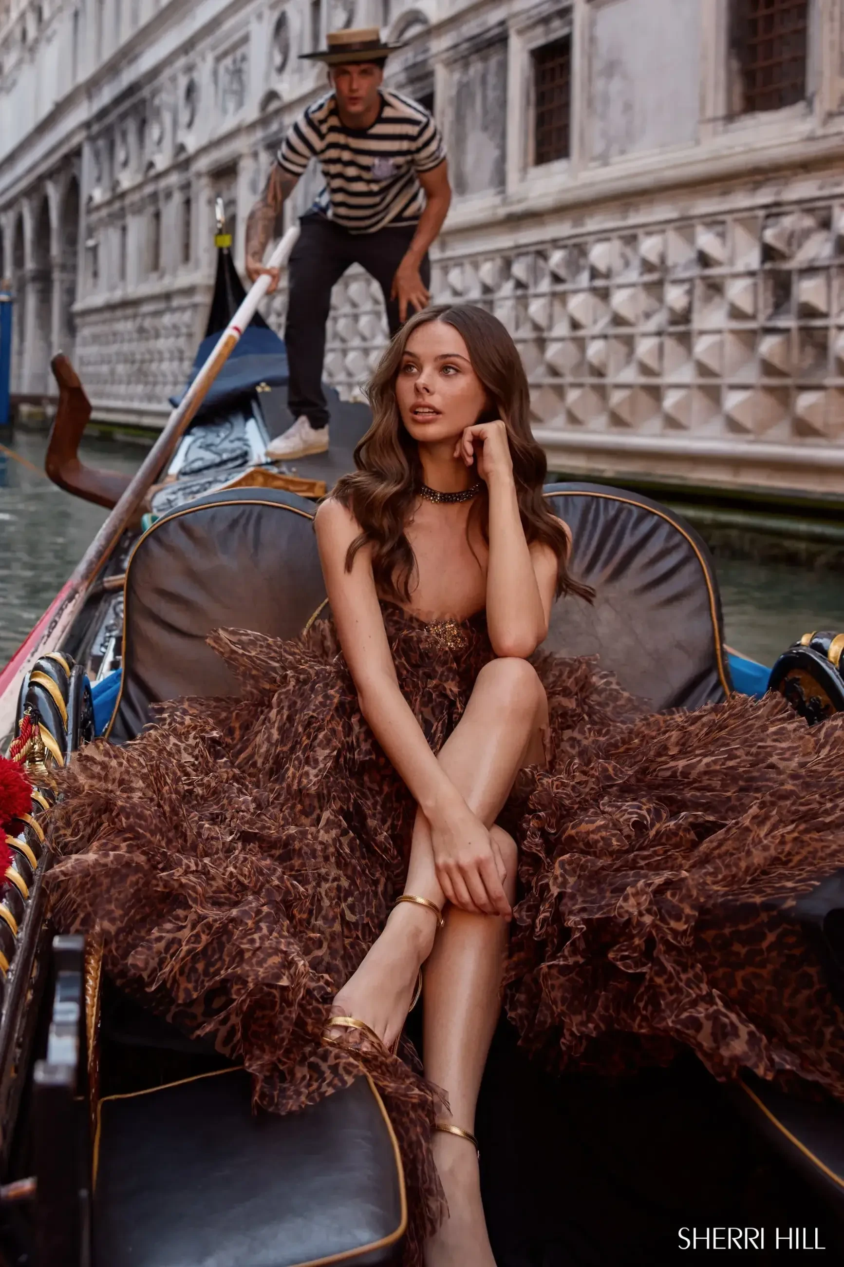 A woman in an elegant brown gown sits in a gondola, posing thoughtfully. A gondolier in a striped shirt rows behind her, with a historic Venetian building in view.