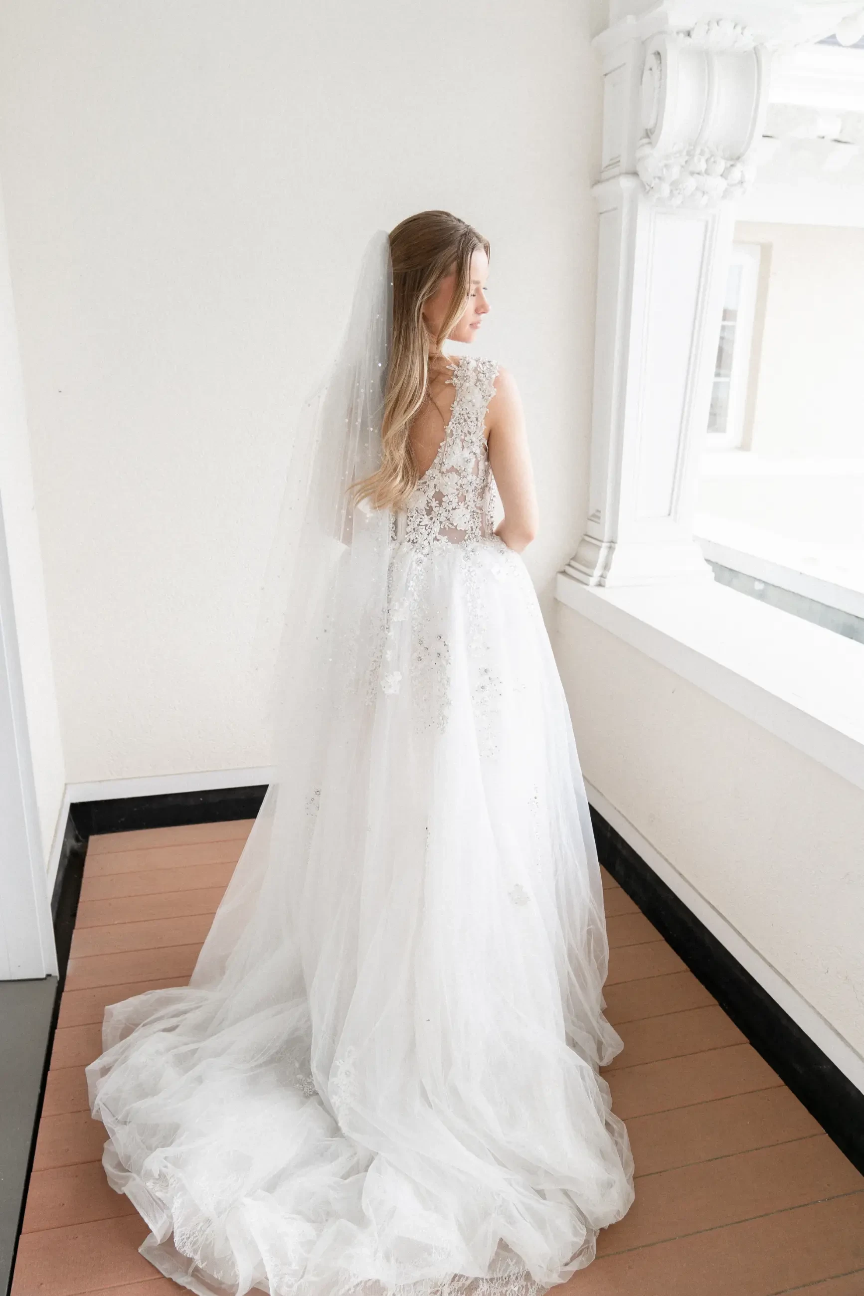 Model is looking over her shoulder and wearing a floor length sleeveless bridal gown with a long train and veil