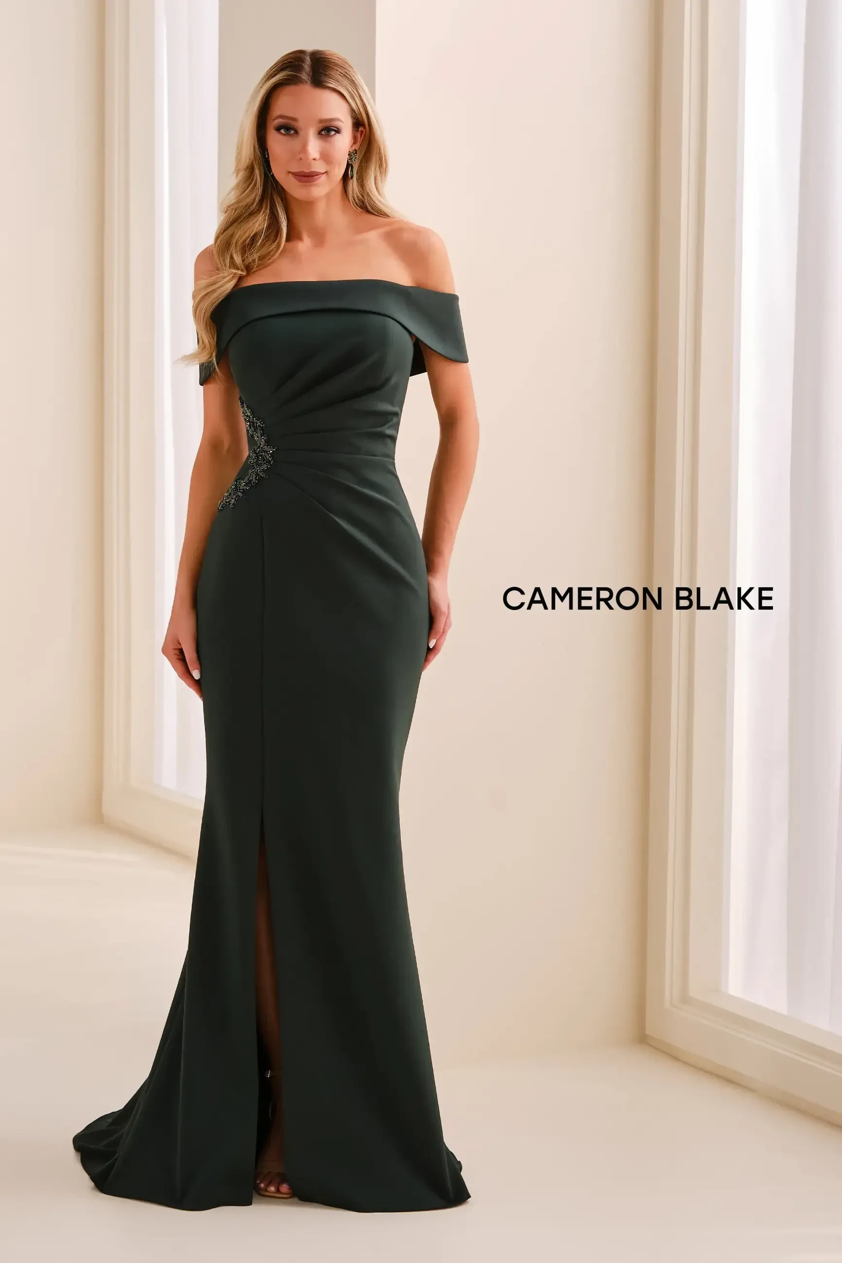 A woman wearing a dark green off-the-shoulder evening gown with a slit, standing in a well-lit room.