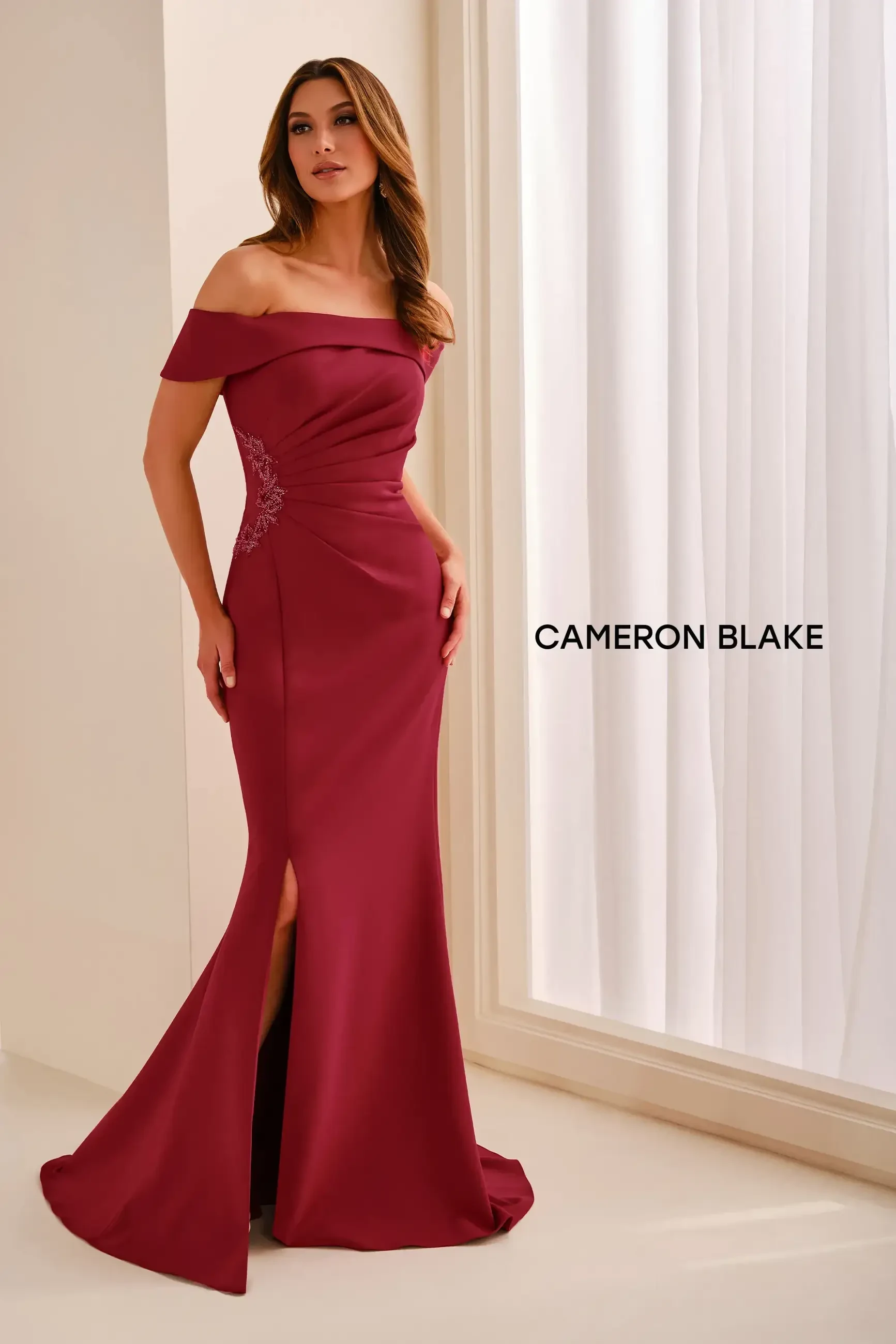 A woman in a burgundy off-the-shoulder dress with a fitted silhouette and a slit, standing gracefully against a light-colored backdrop. The dress features embellishments on one side and the name 
