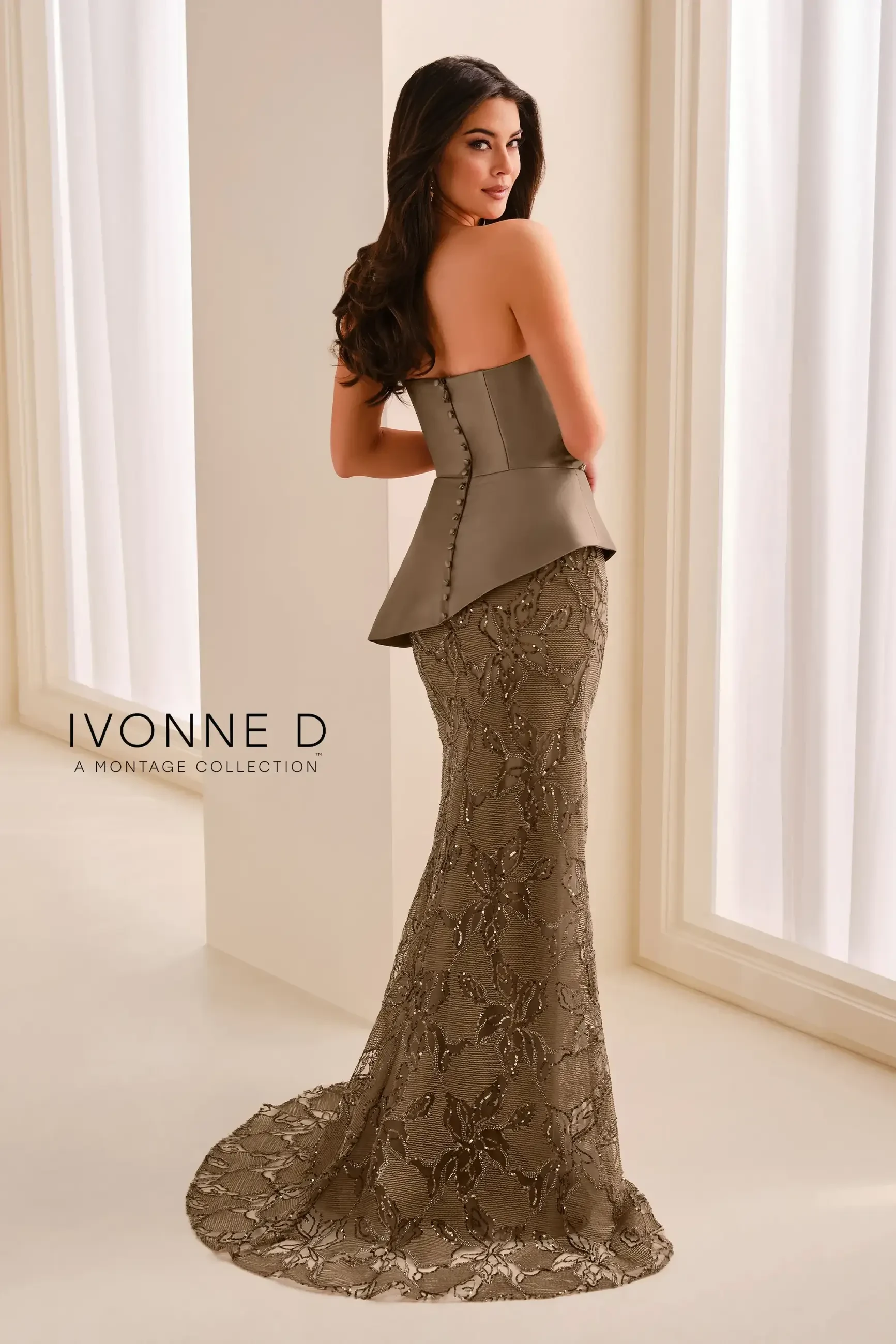 A model wearing a strapless, peplum-style gown with a fitted lace skirt, posing with her back towards the camera in a bright room.