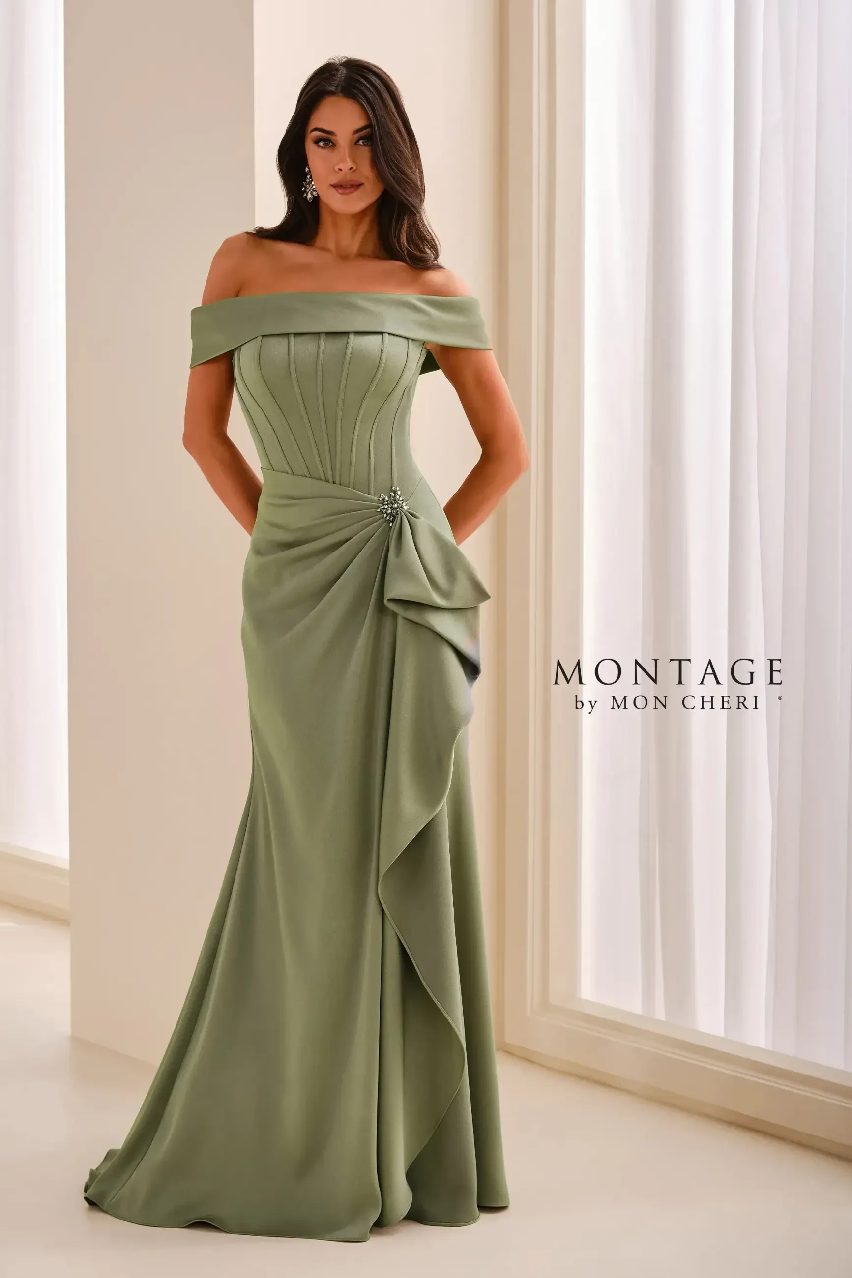 A woman in an elegant off-the-shoulder green gown with a draped design and a side embellishment, standing against a backdrop of soft, natural light.