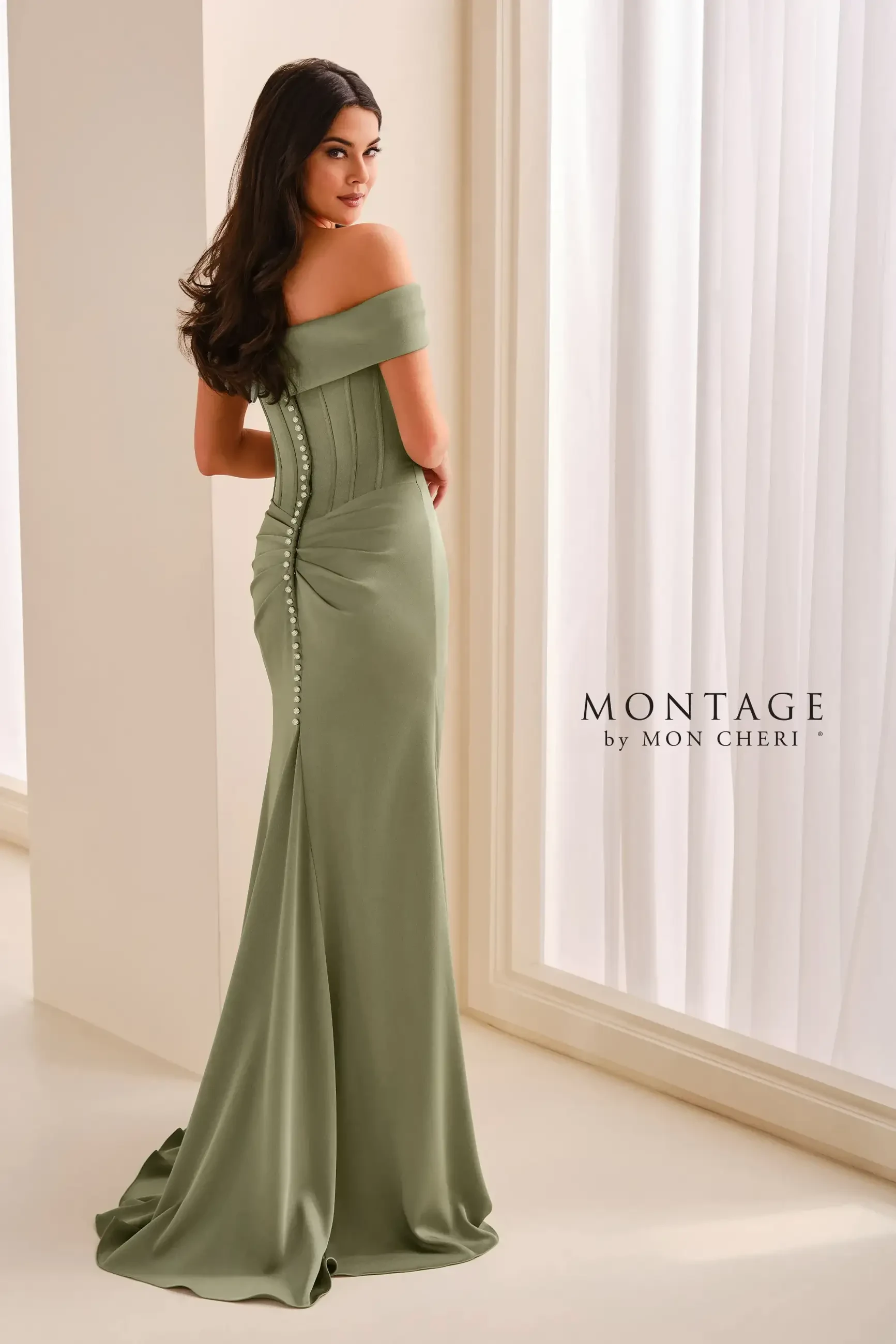 A woman in a green off-the-shoulder gown standing by a window, with her back to the camera, showcasing buttons along the gown's side.